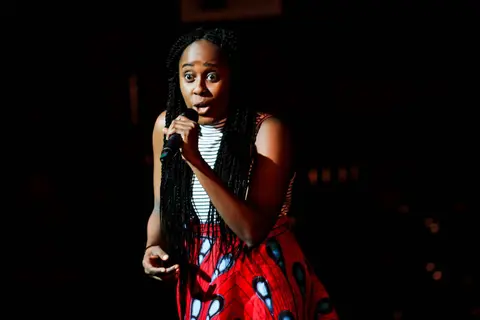 A comedian in a striped top and a red print skirt performs into a microphone under a spotlight.