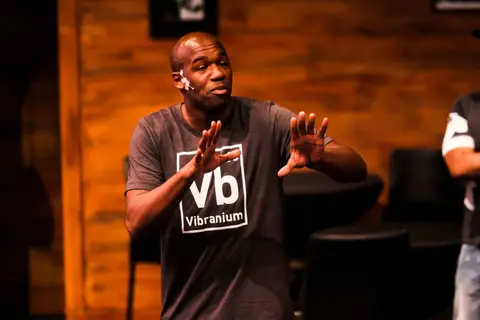 A comedian in a grey T-shirt gestures with both hands mid-joke.
