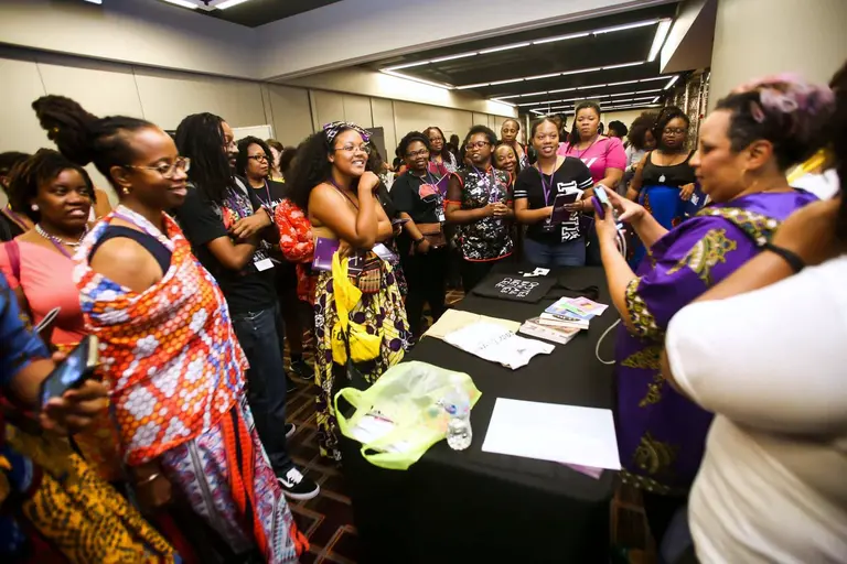 A crowd in bright prints gathers around a vendor's table, phones out, everyone talking.