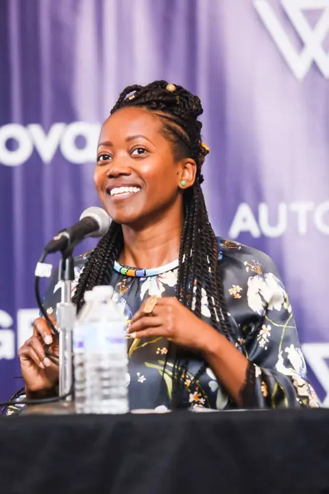 A woman with locs smiles behind a microphone at a panel table.
