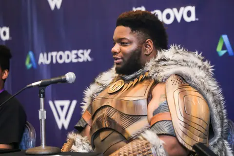 A man in sculpted armour and a fur cape sits at a panel table.