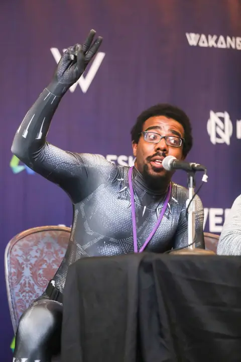 A man in a black bodysuit raises one arm at a panel table.