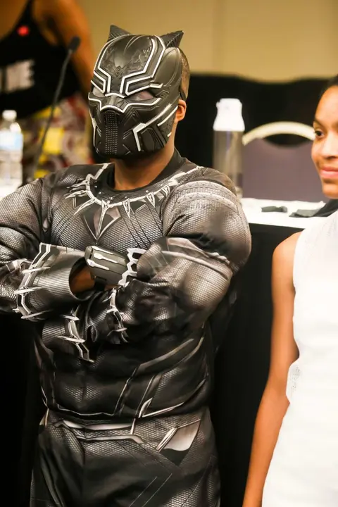 A cosplayer in a full black suit and mask stands with arms folded.