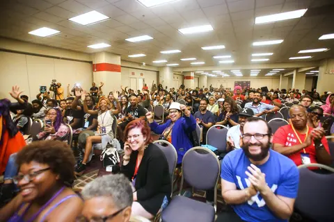 A packed ballroom of people cheering and clapping, arms in the air.