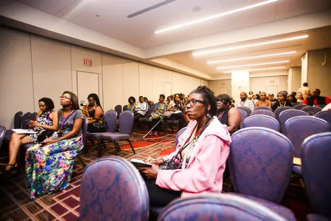 Seated attendees in a hotel meeting room, one in a pink jacket in the foreground.