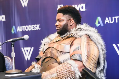 A man in a fur-trimmed armoured costume sits at a panel table with a microphone.