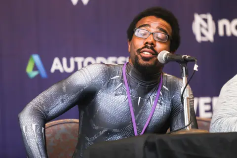 A man in a grey and black superhero suit with glasses speaks into a microphone at a panel.