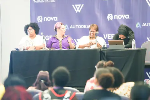 Three women sit at a long panel table in front of the purple backdrop while the audience watches.