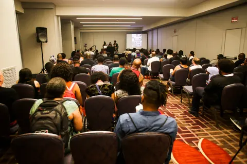 A packed room seen from the back, rows of attendees facing a screen.