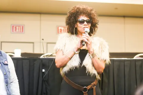 A contestant in sunglasses and a fur vest speaks into a microphone in front of the judges' table.