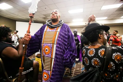A cosplayer in a long purple and gold robe with a staff walks past the cheering crowd.