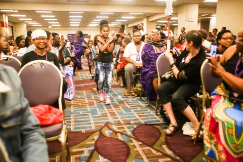 A cosplayer walks between rows of seated, cheering attendees.