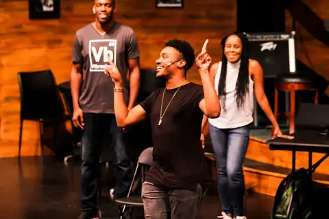 Three performers on a wooden stage, one pointing up mid-joke.