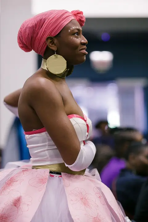 A cosplayer in a pink head wrap, gold hoop earring and a pink and white gown looks up and away.