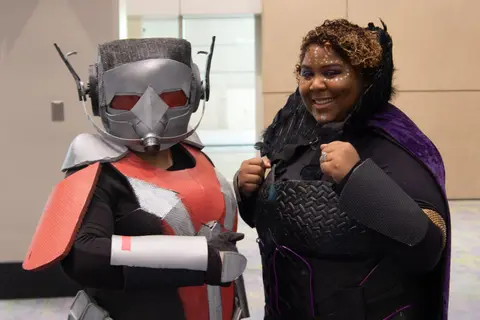 A cosplayer in a red and grey helmet costume next to a smiling woman in a purple cape.