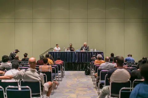 An audience in rows of chairs faces a panel at a draped table in a long room with chartreuse walls.