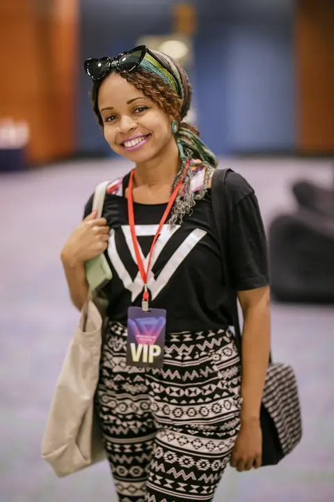 A woman in a head wrap and sunglasses smiles, a VIP badge round her neck and a tote on her shoulder.