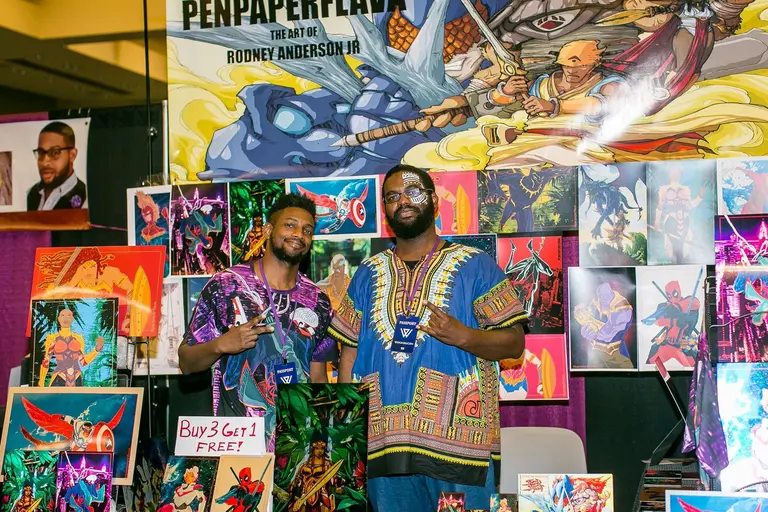 Two artists stand in front of a wall of their own colourful comic prints.