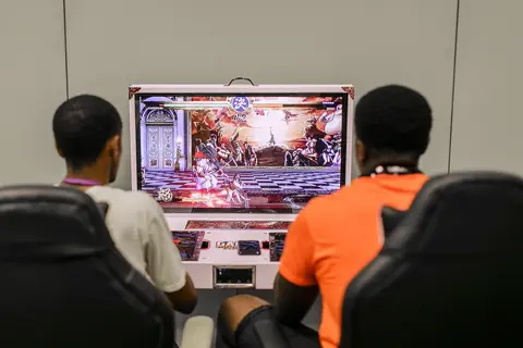 Two players in gaming chairs face an arcade fighting game, seen from behind.