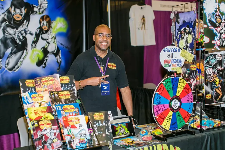A vendor smiles behind a table of comics and a prize wheel.
