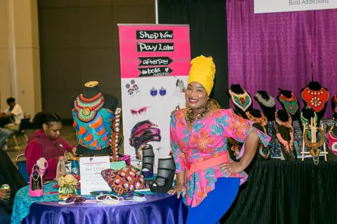 A vendor in a yellow head wrap stands behind displays of beaded jewellery.