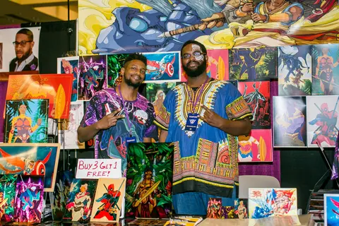 Two men in print shirts stand in front of a wall of colourful paintings.