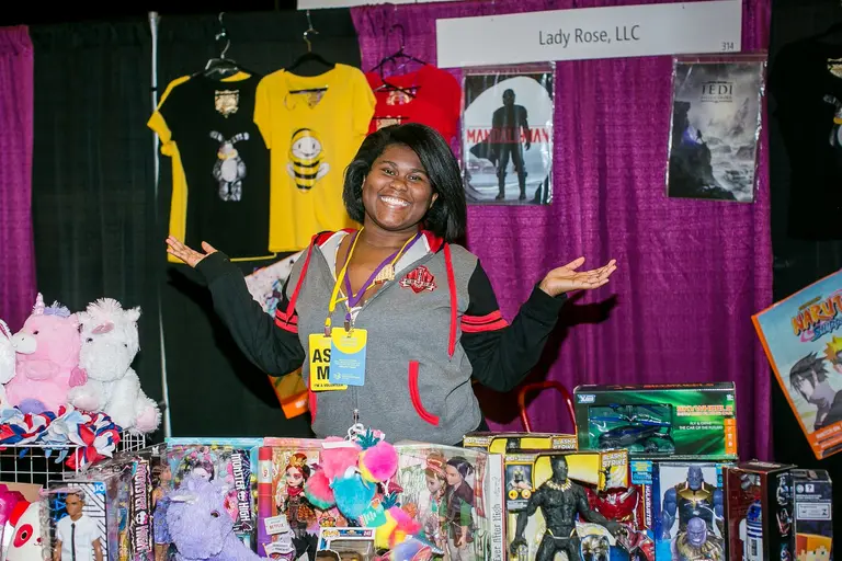 A smiling vendor spreads her arms behind a table of toys and shirts.