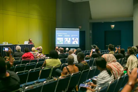 An audience faces a screen and a panel table in a large convention room.