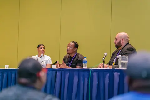 Three panelists speak at a blue draped table, seen over the heads of the audience.