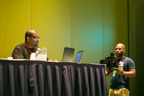 A panelist speaks at a table while a man films with a camera rig.