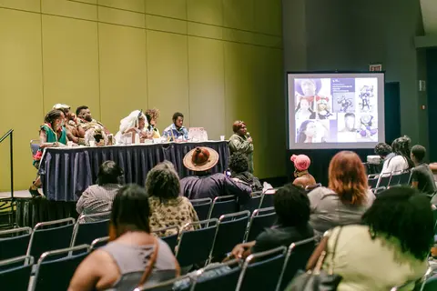 Panelists in costume sit at a draped table under a projected slide.
