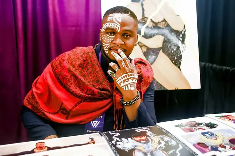 An artist in white face paint and a red shawl sits behind his table of prints.