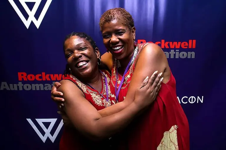 Two women in red hug and laugh at the blue W backdrop.